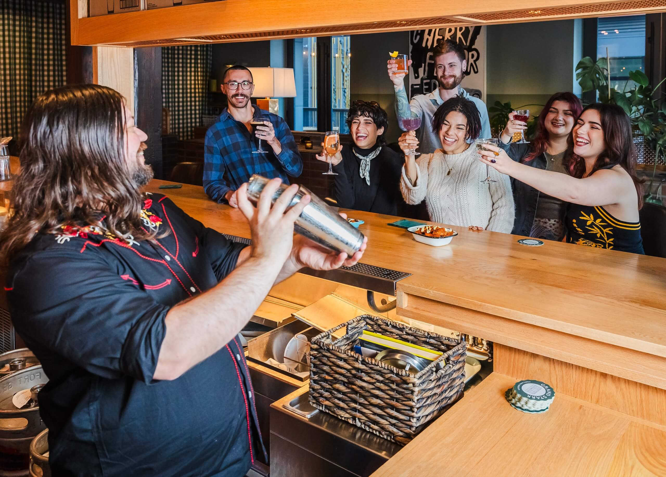A bartender shaking a cocktail shaker while a group of people cheer and hold up their drinks in the background.
