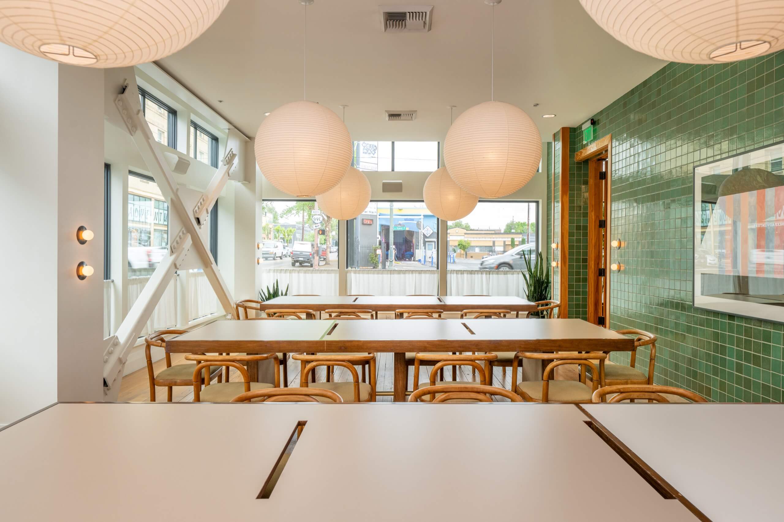 A brightly lit dining area with large tables and wooden chairs, featuring paper lanterns and a green tile wall.