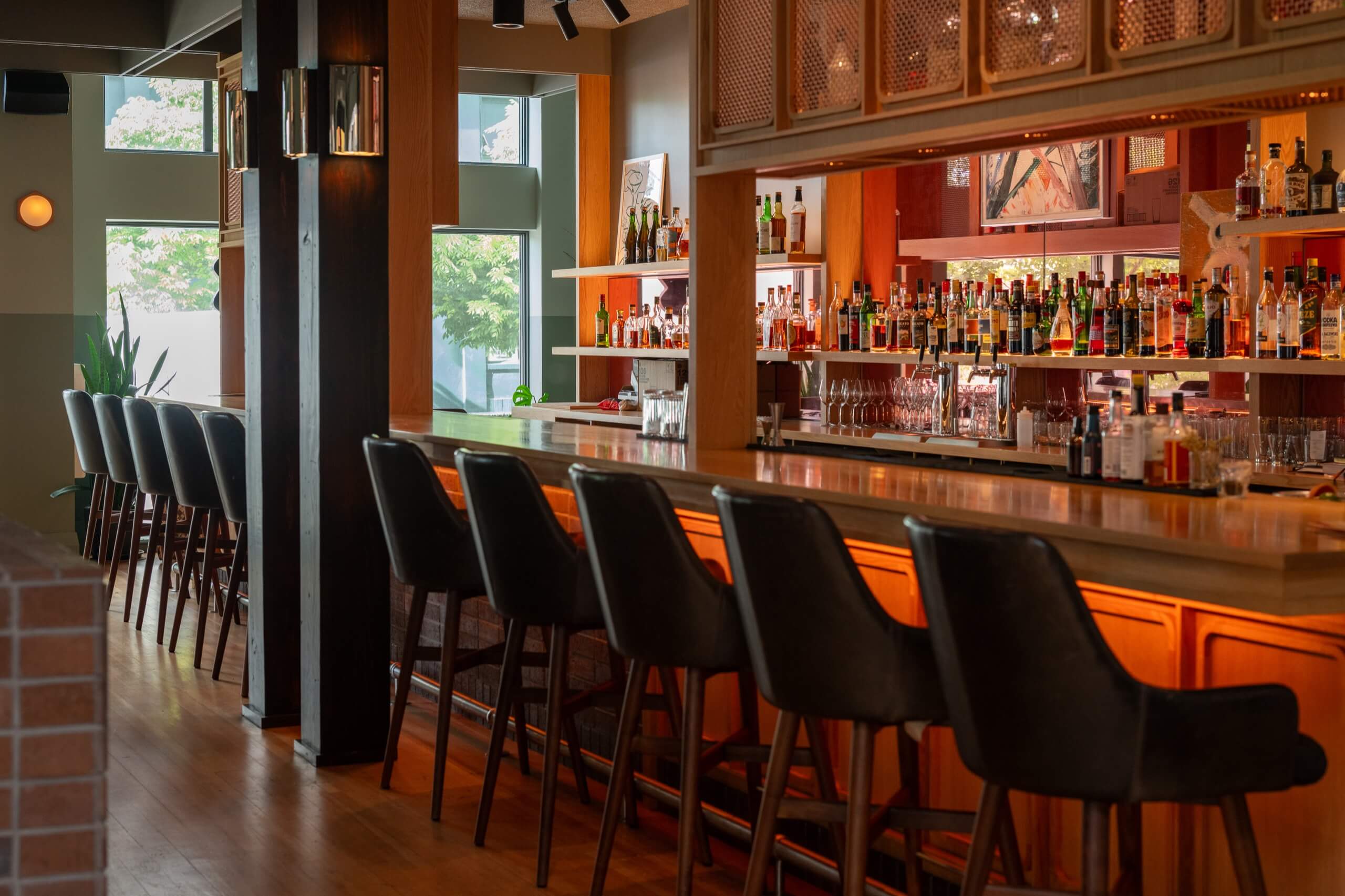 A dimly lit bar with a long counter and several stools, shelves of bottles in the background.