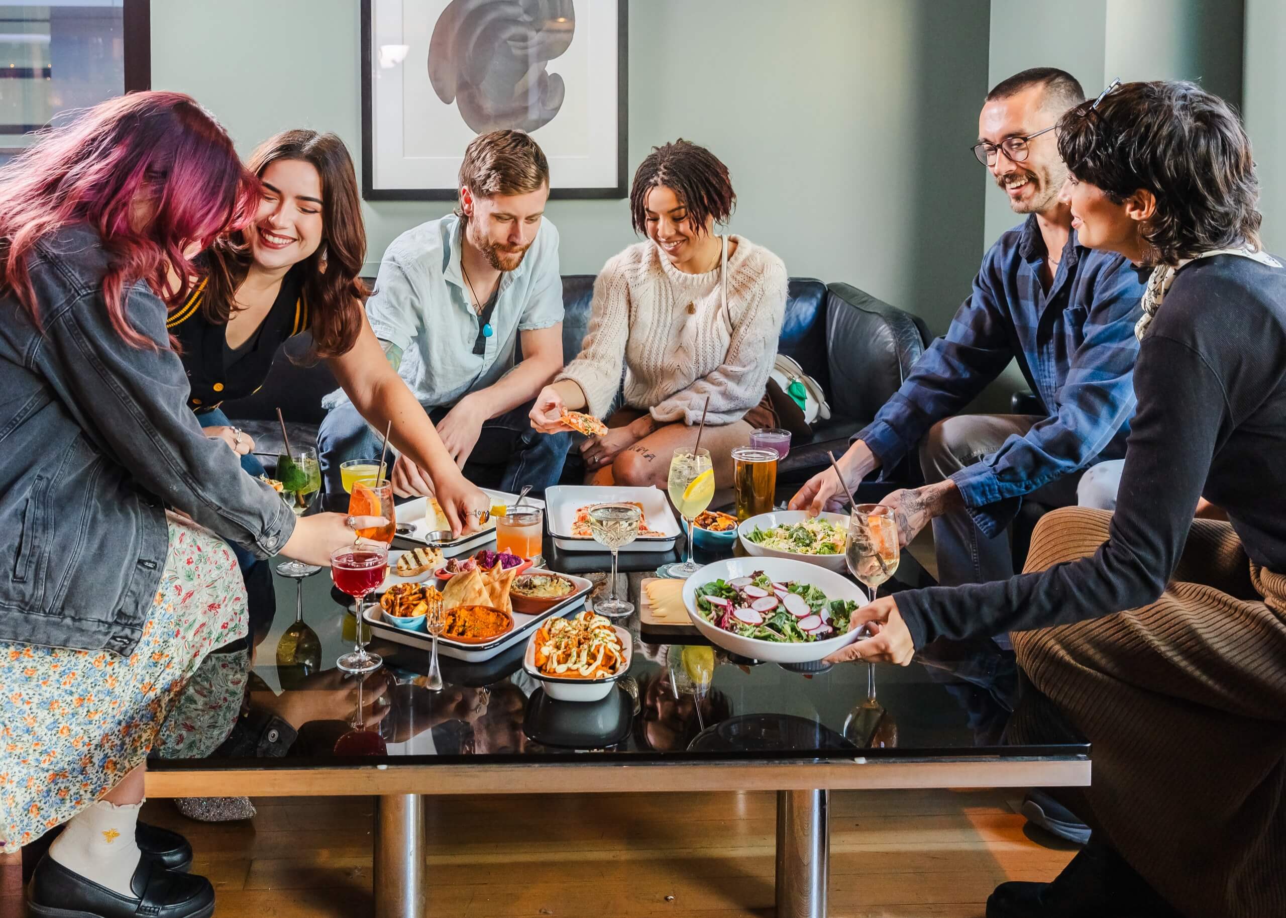 A group of six people sitting around a table with food and drinks in a lounge area.