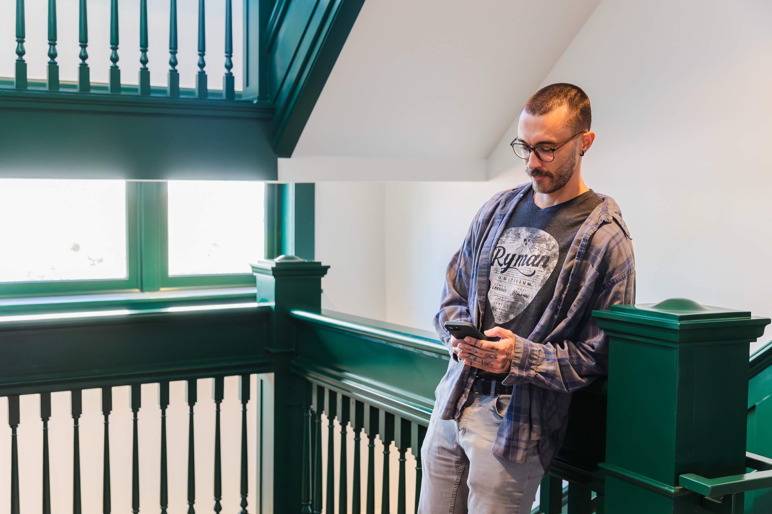 A man stands on a staircase with green balustrades and handrails, looking at his phone.
